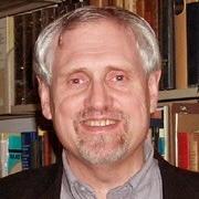 Keith Dennis, with goatee and mustache, standing in front of book case.