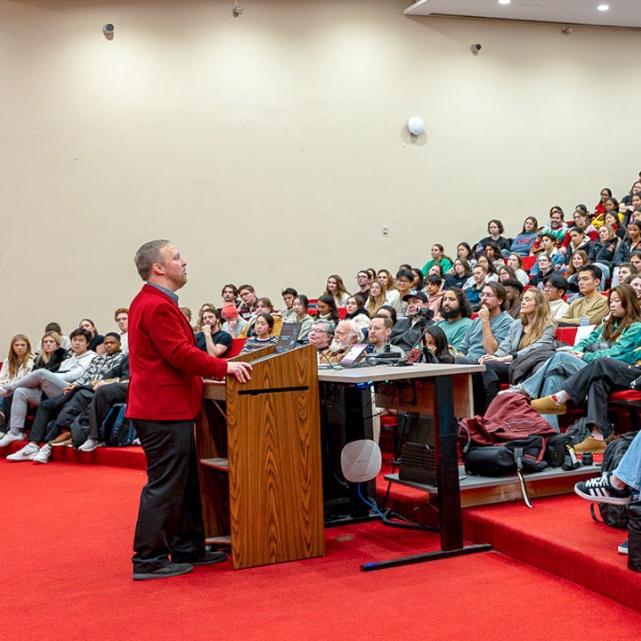 Person wearing a red jacket speaks at a podium, to an auditorium full of students