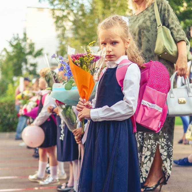 		Girl wearing a backpack and holding flowers, in a line of other students
	