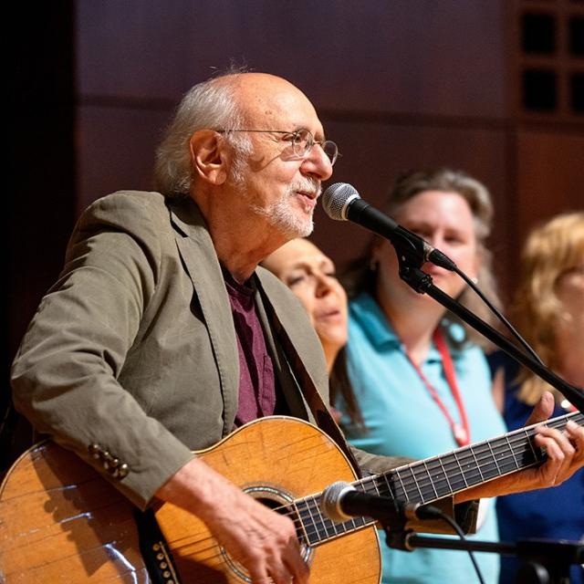 		Peter Yarrow with guitar, tan jacket, white hair just at the back of his head, white beard and glasses in front of a microphone with three women and a child standing next to him
	