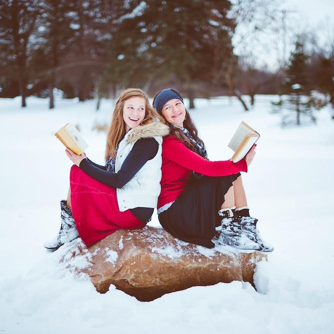 		Two people wearing fashionable red, white and black winter clothing sit back to back on a large rock, each holding a book. They are surrounded by snow
	