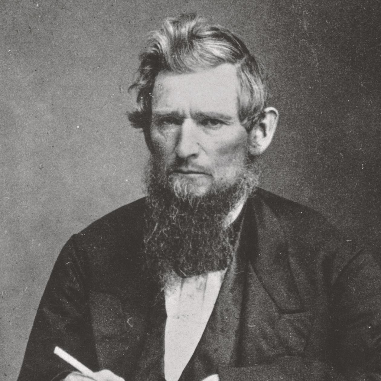 Black and white historic photo of Ezra Cornell, frowning and holding a pen. 