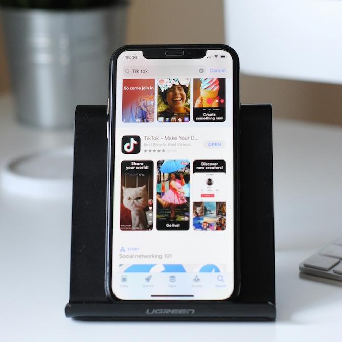 		smart phone on a stand on a desk, showing TikTok home page
	