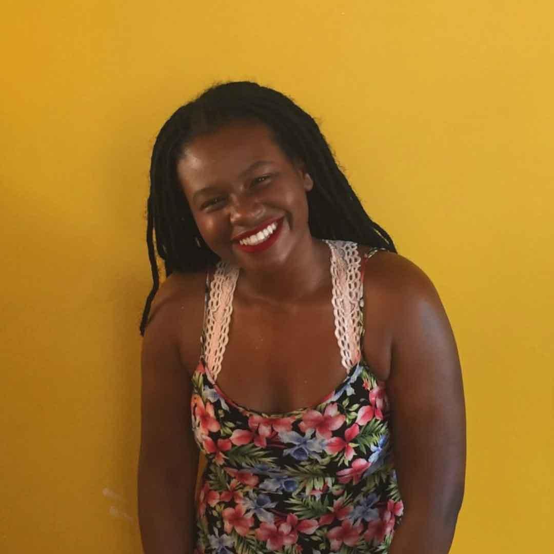 Brittani Samuel, head tilted to the right, smiling broadly, with long hair in small tight braids, wearing a flowered sleeveless dress