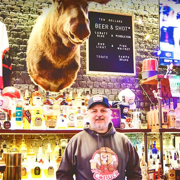 		Person standing behind a bar decorated with a hockey jersey, a beer & shot list and a mounted moose head. Lots of neon lights and two illuminated shelves of liquor bottles
	