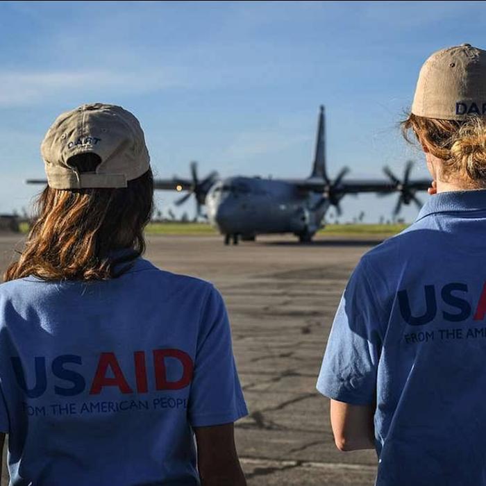 		Two people on a tarmack facing a plane. They're wearing shirts with "USAID" written across the back
	