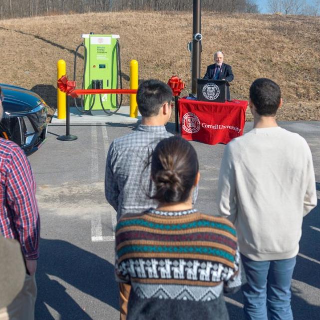 		Sevearl people stand in a parking lot, listening to a speaker who stands at a makeshift podium next to a large green machine
	