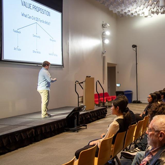 		Person speaking on a small stage with a large audience watching
	