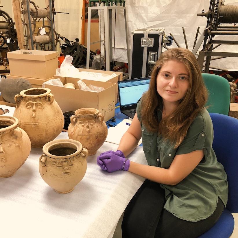 		Person wearing laytex gloves sitting at a table with four ancient artifacts: pots
	