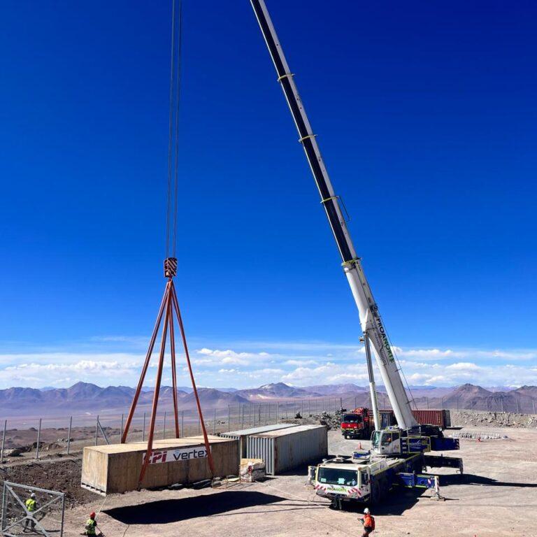 Tall crane lowering a huge rectangular crate onto the ground with mountains in the background.