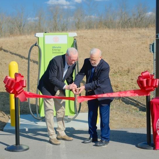 		Two people cutting a red ribbon ceremonially. They are outdoors
	