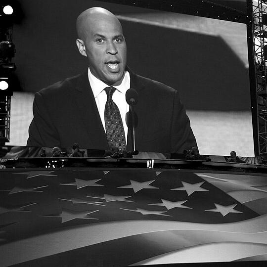 Corey Booker in suit and tie on a large screen with a huge American flag in front of it,