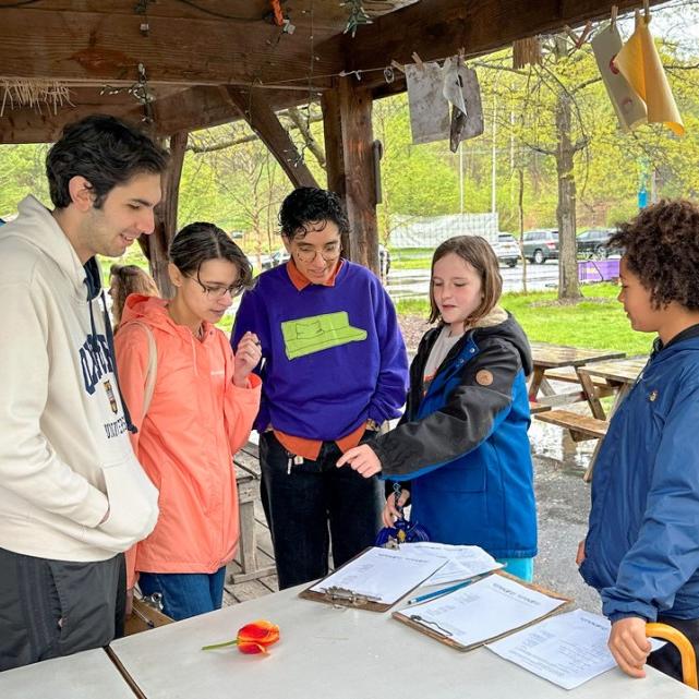 		People cluster together in an outdoor shelter, looking at papers and flowers
	