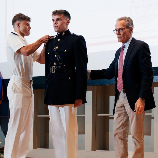 		People on a stage; one in military uniform stands at attention
	