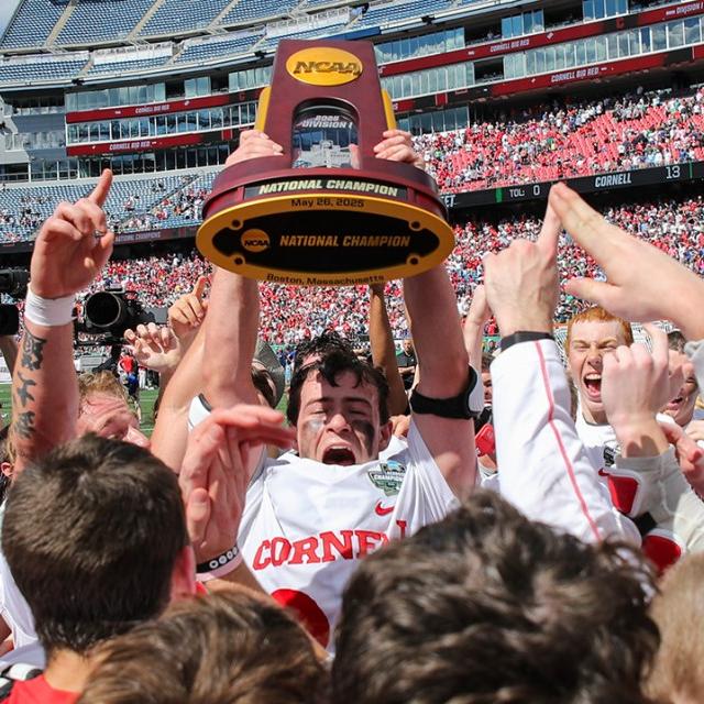 		A lacross player in white and red uniform holds a trophy aloft; he's surrounded by celebrating teammates
	