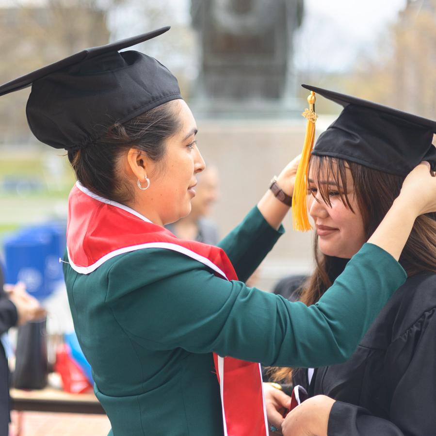 		One person wearing graduation cap helps another with hers
	