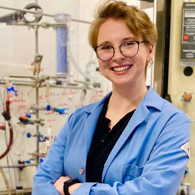 		Beth Ryan with big glasses, a smile and a lab coat standing in front of a white board with equations and a lab frame with hoses and things hanging on it
	