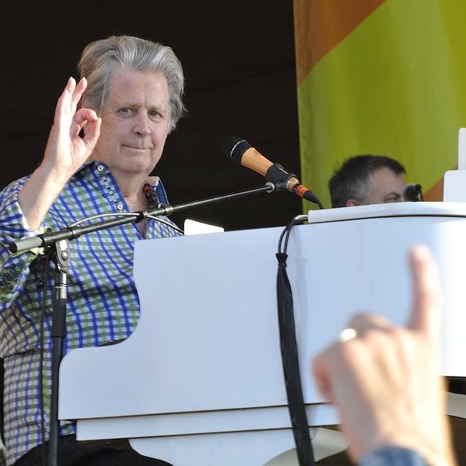 		Person sitting at a white piano on a stage, giving an "okay" sign with hand
	