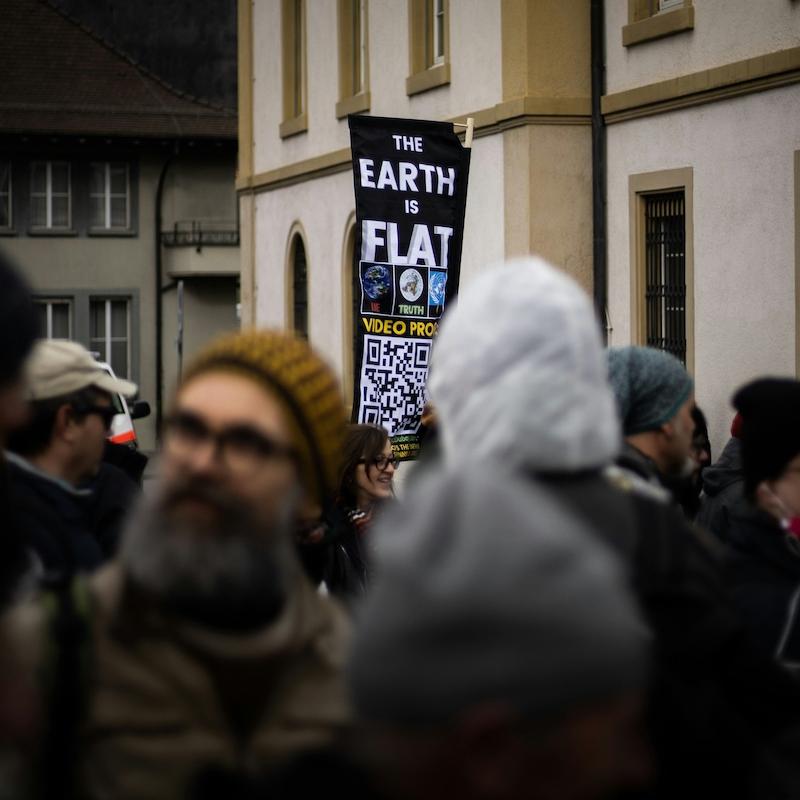 A sign sticks out from a crowd of people, saying "the Earth is flat)