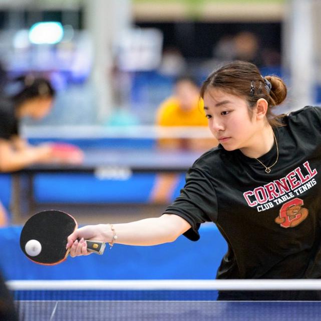 		Person in black Cornell t-shirt reaches gracefully to hit a small white ball with a paddle
	