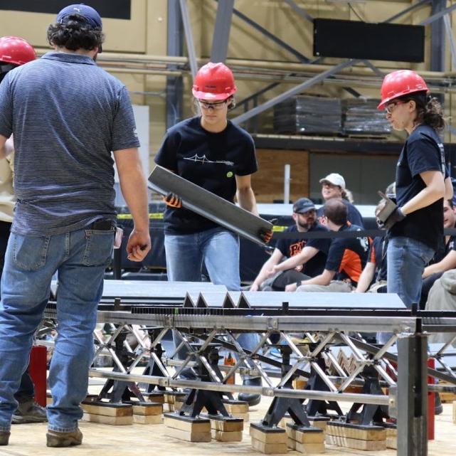 People in red hard hats construct a structure on the floor of a large indoor space