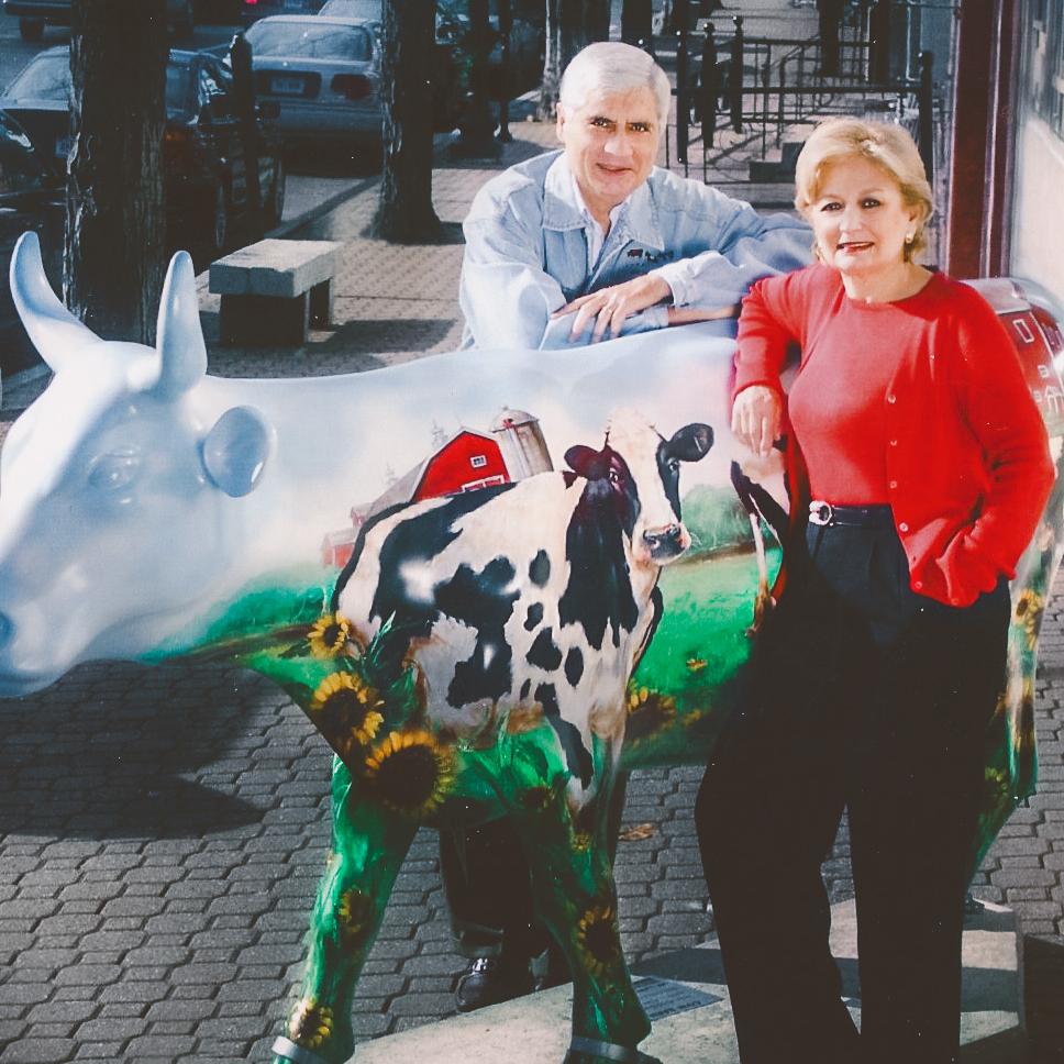 		A white haired man and a woman in bright red shirt and sweater stand leaning against a cow with a painting of a cow on it, with a barn in the back and sunflowers and grass on its legs.
	