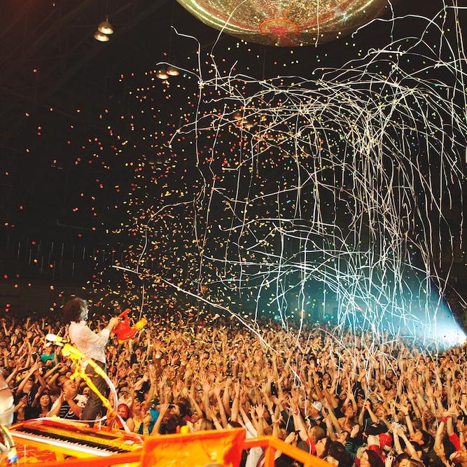 A crowd raises their hands with excitement as a band plays on a stage (barely in the image) and confetti rains down from above