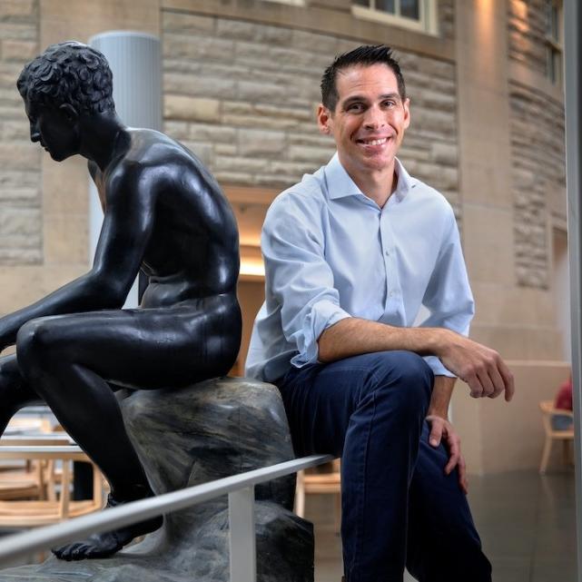 		Person sitting casually back to back with a statue of a seated human figure carved from black stone
	
