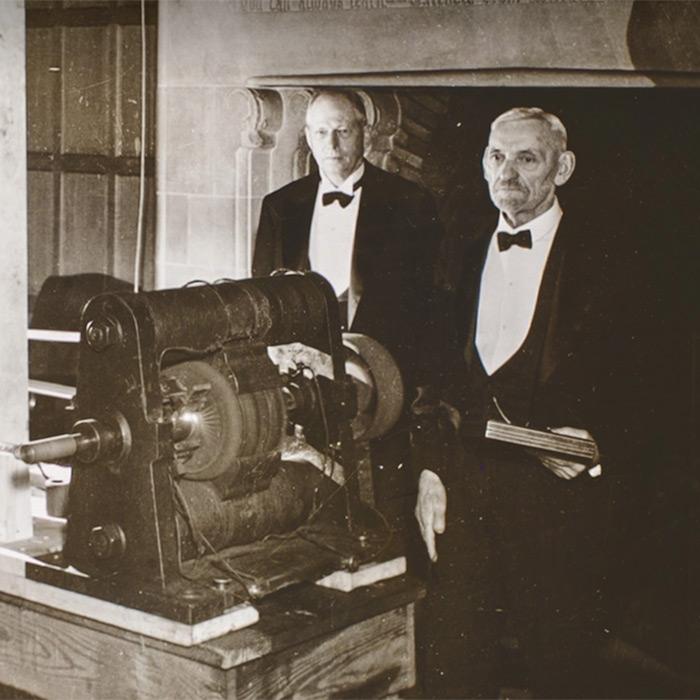 Historic black and white photo of three people in evening coats standing around a machine