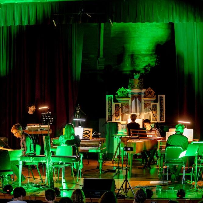 		A stage bathed in green light: several people play on keyboard instruments, including standing sythesizers, pianos, and an organ
	