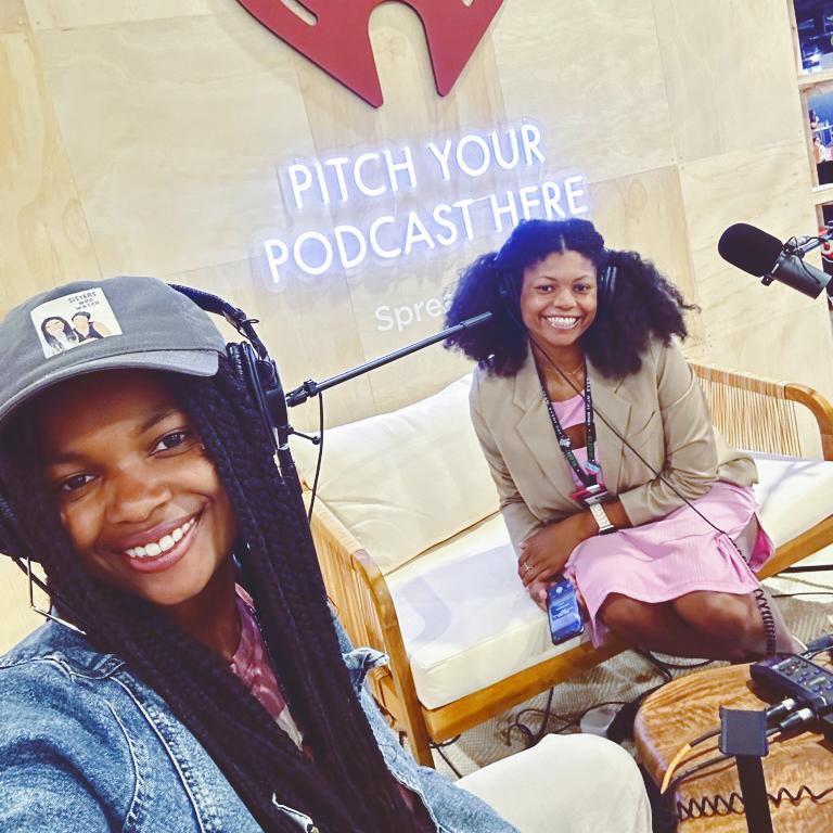 		Two people wearing headphones and recording equipment under a neon sign: Pitch your podcast here
	