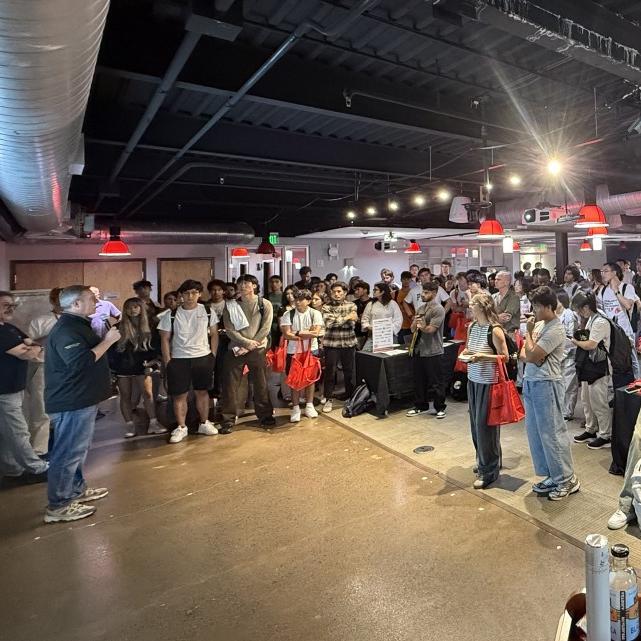 Lots of students standing in a big space with bright lights overhead facing someone with a microphone.