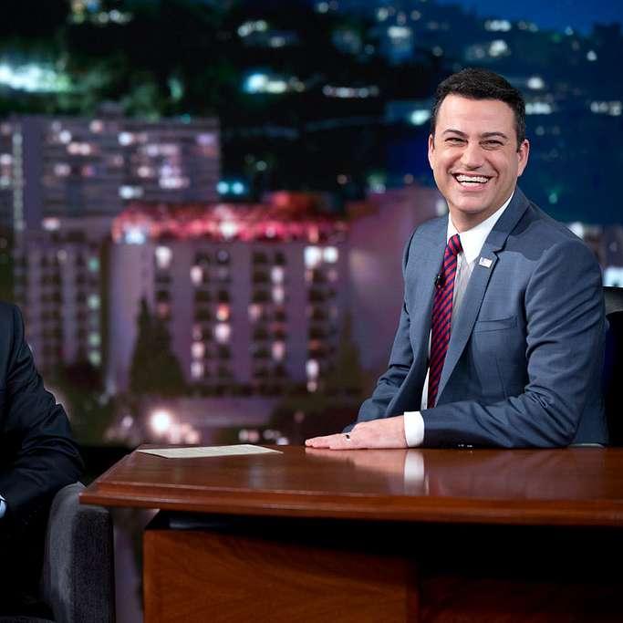 Two people sit at a desk on the set of a late night TV chat show