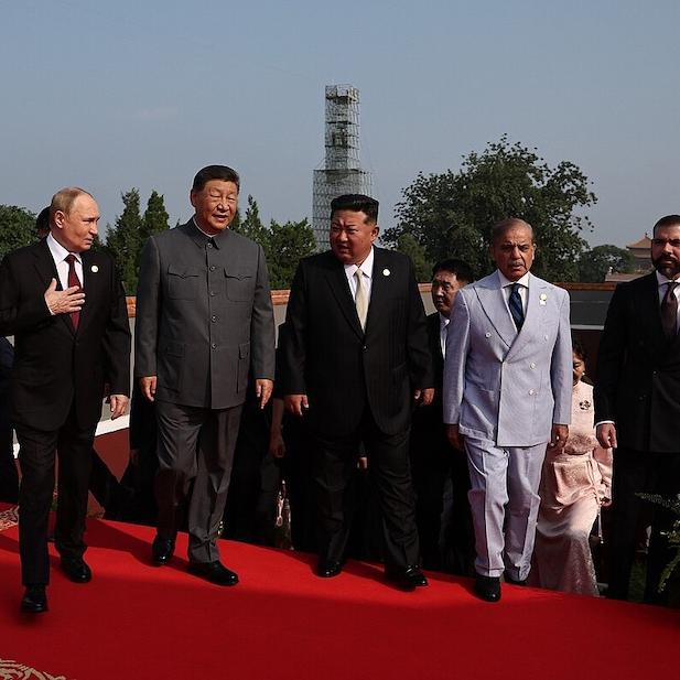 Six people in suits walk abreast up a red carpet, conversing
