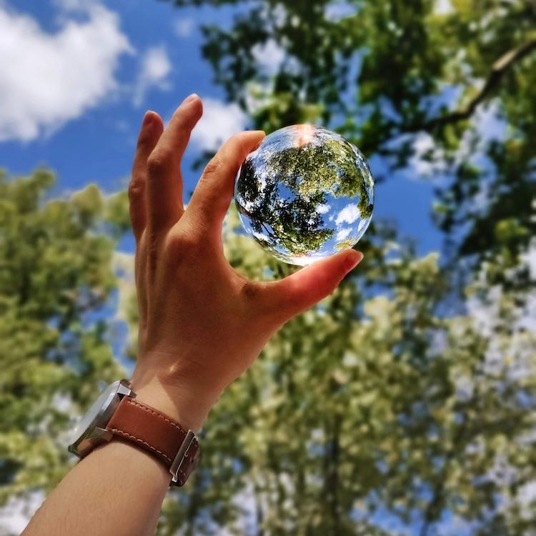 Hand holding a glass sphere up toward the sky so it reflects trees and clouds, making the ball look like a tiny planet Earth
