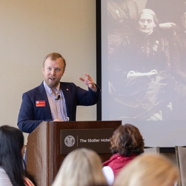 Person speaking at a lecturn with a black and white photograph displayed behind