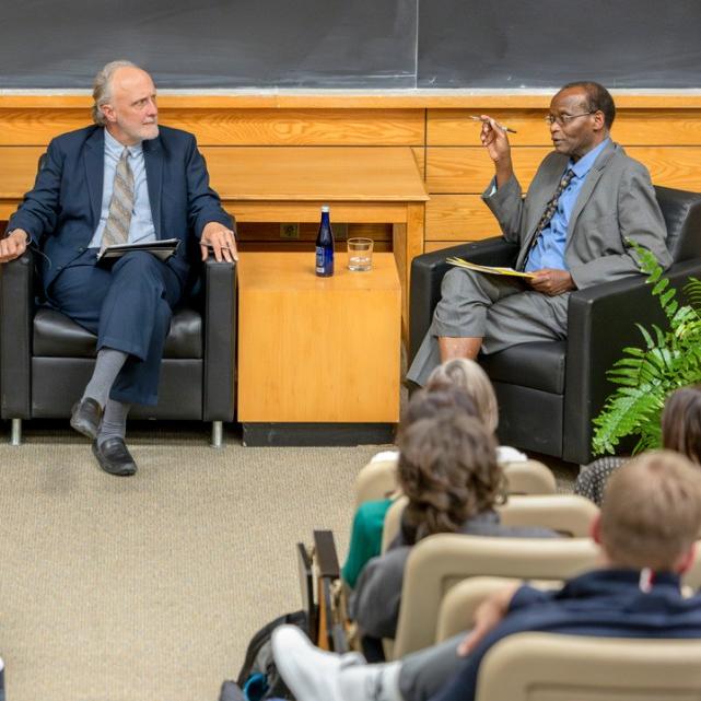 		Three people sit in chairs in front of audience during a debate
	