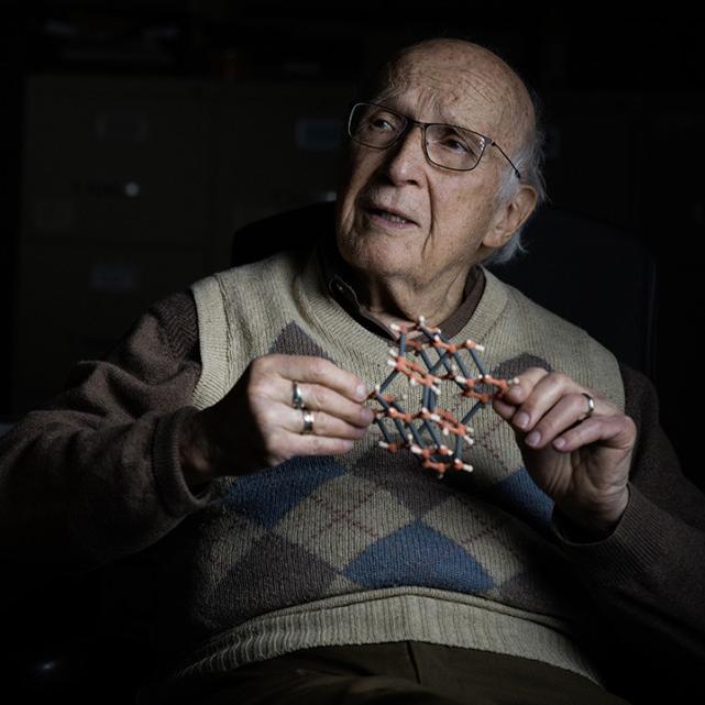 		Artistic portrait of a person in argyle pattern sweater vest holds a small model of a molecule and looks pensive
	