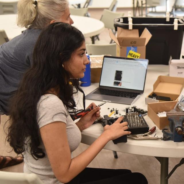 		Two people sit at a table loaded with equipment, assembling a robot
	