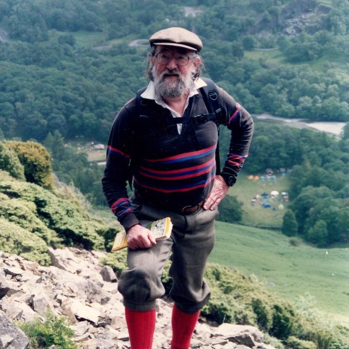 Person wearing a cap, sweater, wool hiking pants and sturdy boots, poses on rocky ground of a very steep hill
