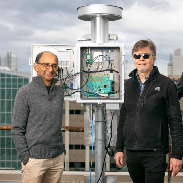 		Three people stand next to a silver device, about a foot taller than the people, made of a post, a box with electronics inside, and a circular top
	