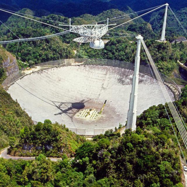 Large concrete dish surrounded by three poles and wires; a mechanism is suspended over the dish. the whole thing, a telescope, is surrounded by lush trees