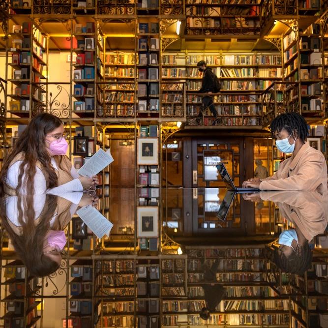 Two people sitting at a table, reading in a room full of books on shelves; the books are reflected on the surface of the table, creating a sense of immersion