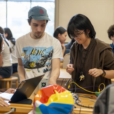 		About five students cluster around a table full of equipment in an introductory physics lab
	