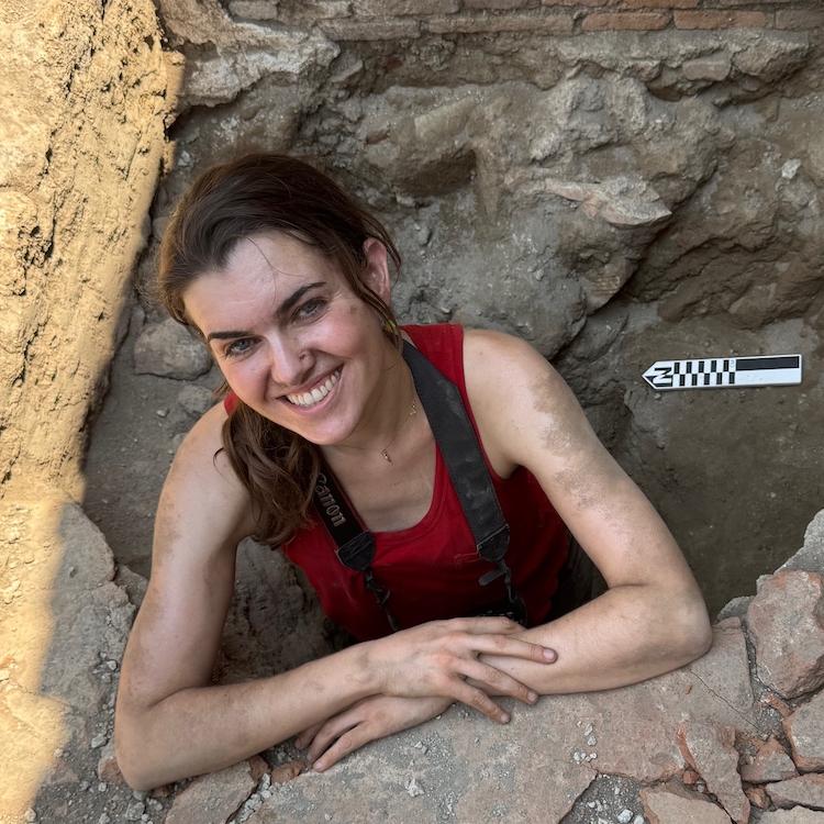 		Person with dust on face and arms, smiling while standing in a hole in the ground, an archaeological dig site
	