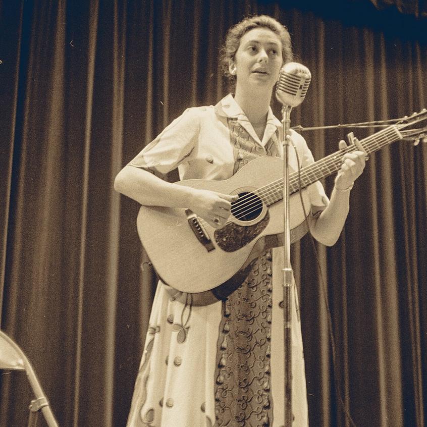 		Black and white historic photo of a person playing a guitar and singing into a microphone
	