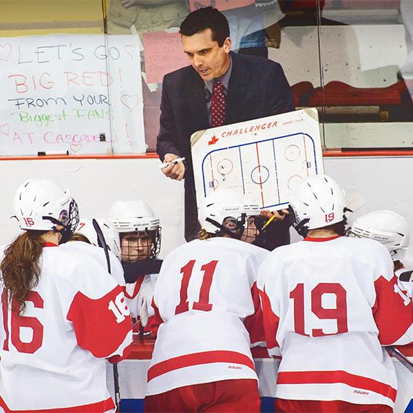 A coach with a white board in front of several hockey players in red and white jerseys