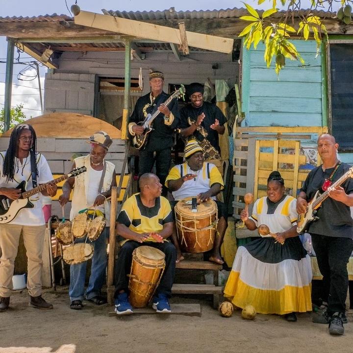 		Several people standing around and sitting on porch steps in the shade, playing drums, guitars and maracas
	
