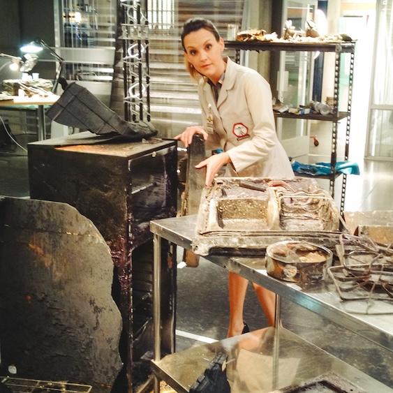 Actor on the set of a lab, wearing a chic trench coat, among a collection of burned or corrupted objects