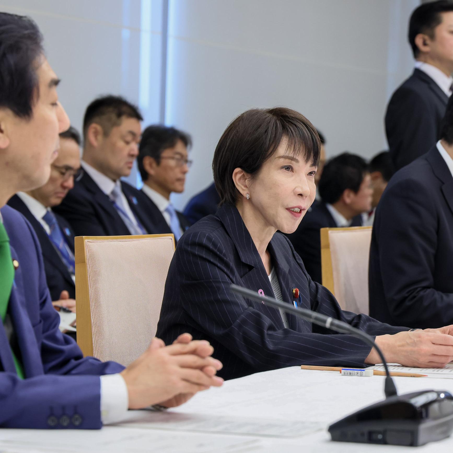 		Sanae Takaichi in dark suit jacket sits at a table with microphones between two men, and more men sitting in the room behind her
	
